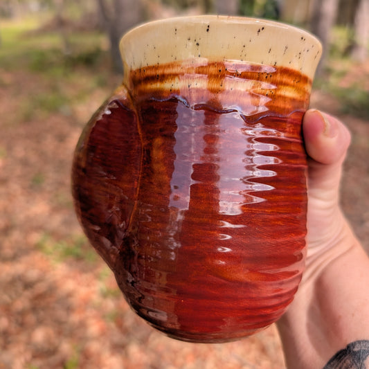 Hand holding a ceramic mug with a red-brown glaze against a blurred natural background