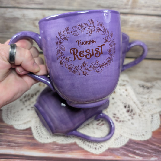 Purple mug with 'Fucking Resist' text held by a hand on a wooden surface.