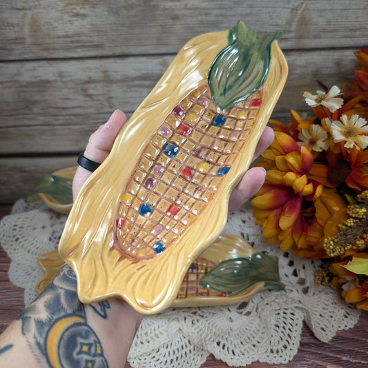 Long thin dishes designed to hold corn on the cob, painted to look like Indian corn on a wooden surface with flowers off to the side.