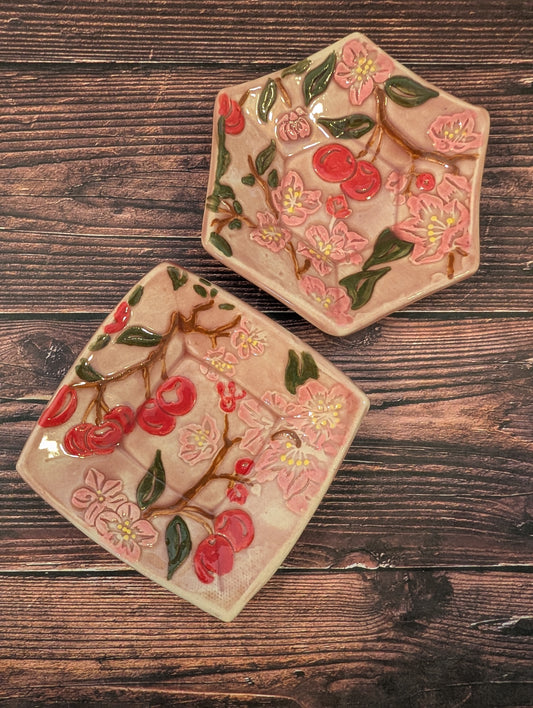Two trinket dishes, one square and one hexagon. Both are light pink with detail painted cherries and cherry blossoms.