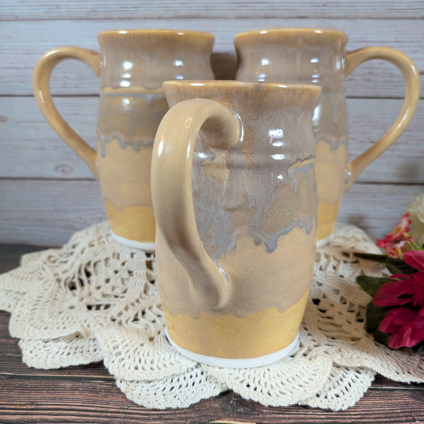 Three large mugs in varying shades of yellow, on a lace doily on a wooden surface. There are red flowers off to the right.