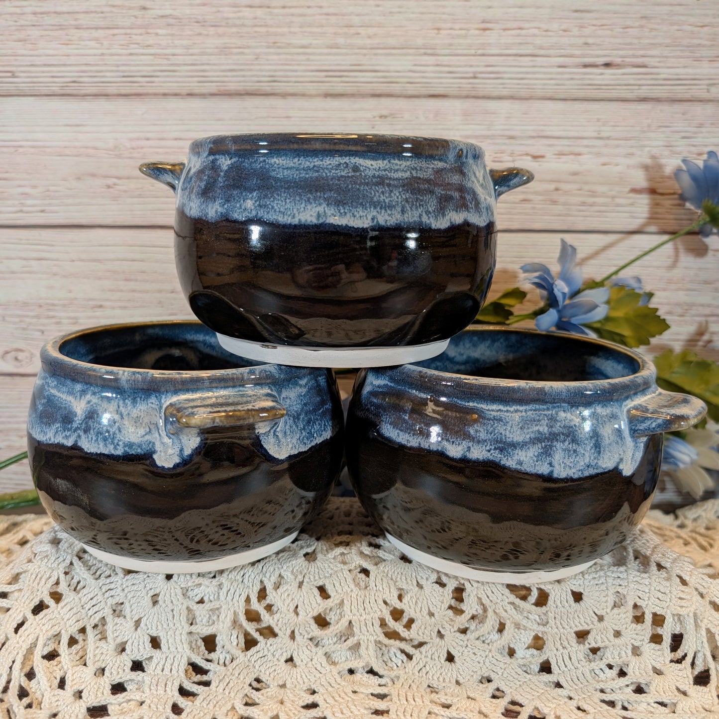 Three small glazed ceramic pots with side handles, stacked with one on top of two, on a white lace doily against a light wood plank background with blue flowers.