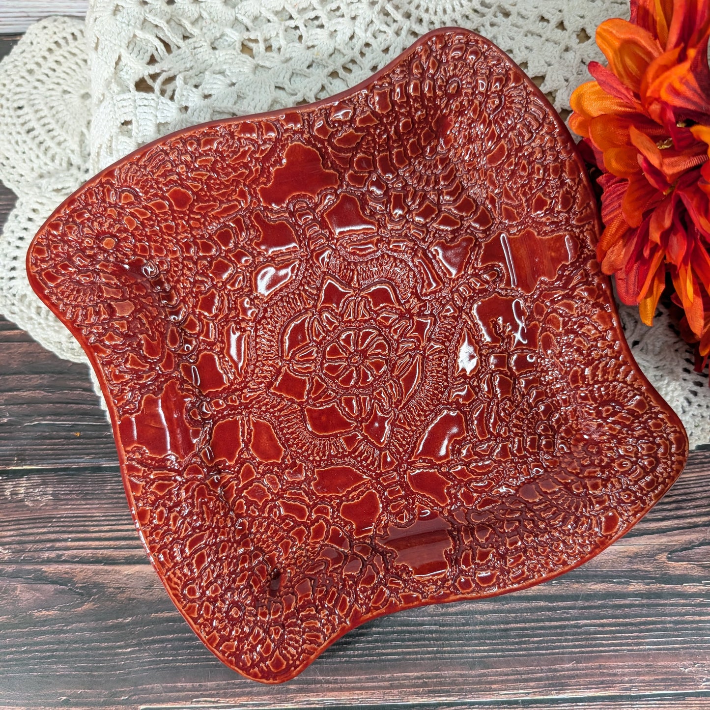 A square ceramic plate with scalloped edges, featuring a glossy, raised floral and geometric pattern in a deep reddish-brown glaze. The platter is styled on a dark wood surface with a white crocheted lace cloth and a red-orange flower in the background.