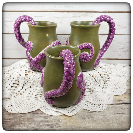 A set of three tentacle mugs with green bodies and purple tentacle handles, displayed on a white doily.
