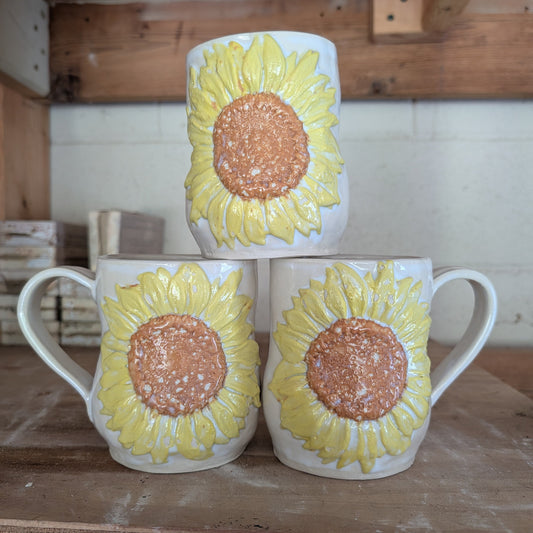A stack of three mugs, white, with sunflowers. 