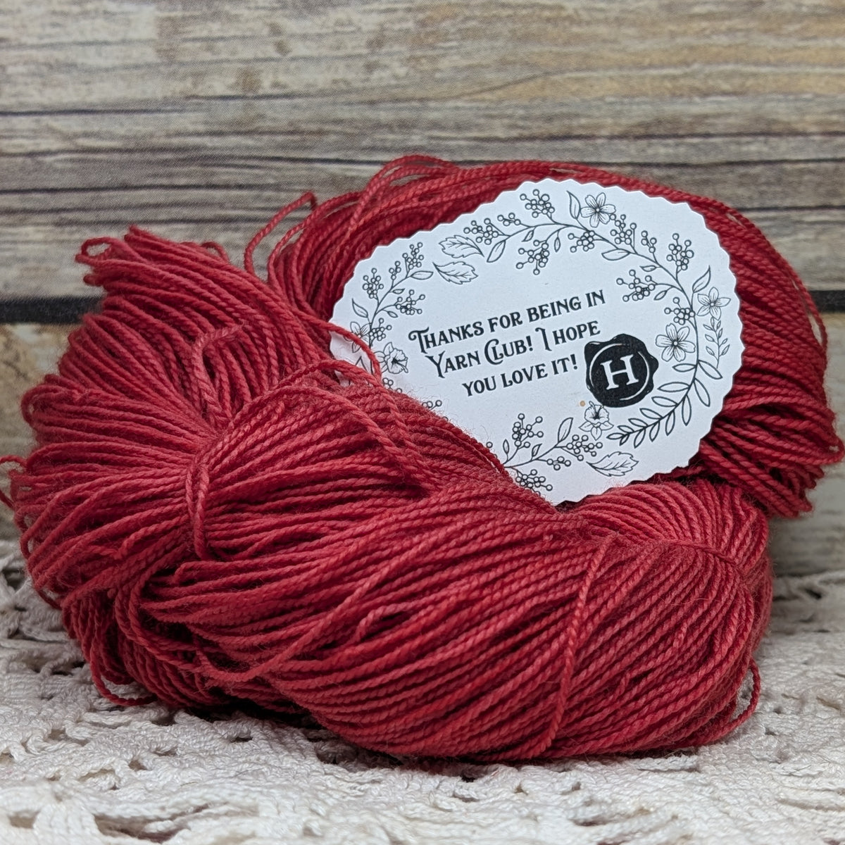 A skein of deep red yarn with a white paper thank-you label, placed on a rustic wooden surface.