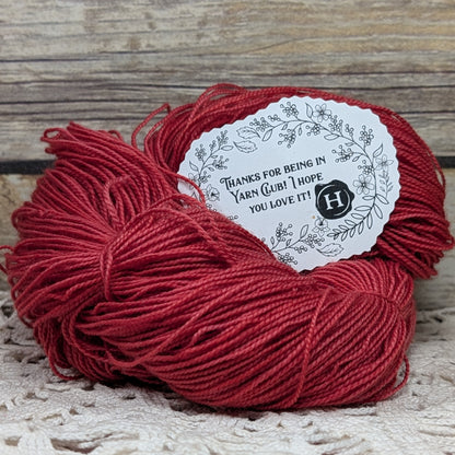 A skein of deep red yarn with a white paper thank-you label, placed on a rustic wooden surface.