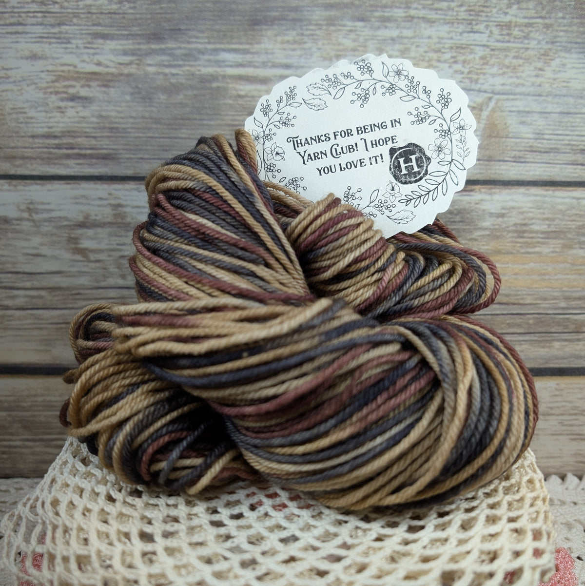 A skein of variegated yarn with brown, beige, and burgundy stripes, displayed on a white mesh surface with a paper tag attached against a rustic wood background.