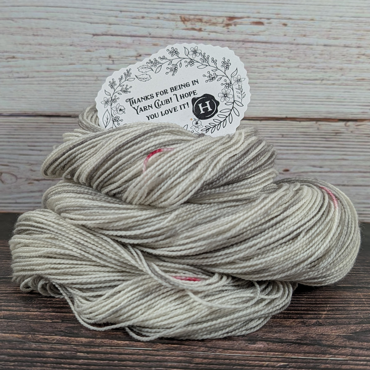 A skein of variegated yarn in light taupe-gray with subtle pink and cream variegation, displayed on a dark wooden surface with a rustic whitewashed wood background. A paper tag is tucked into the top of the yarn.