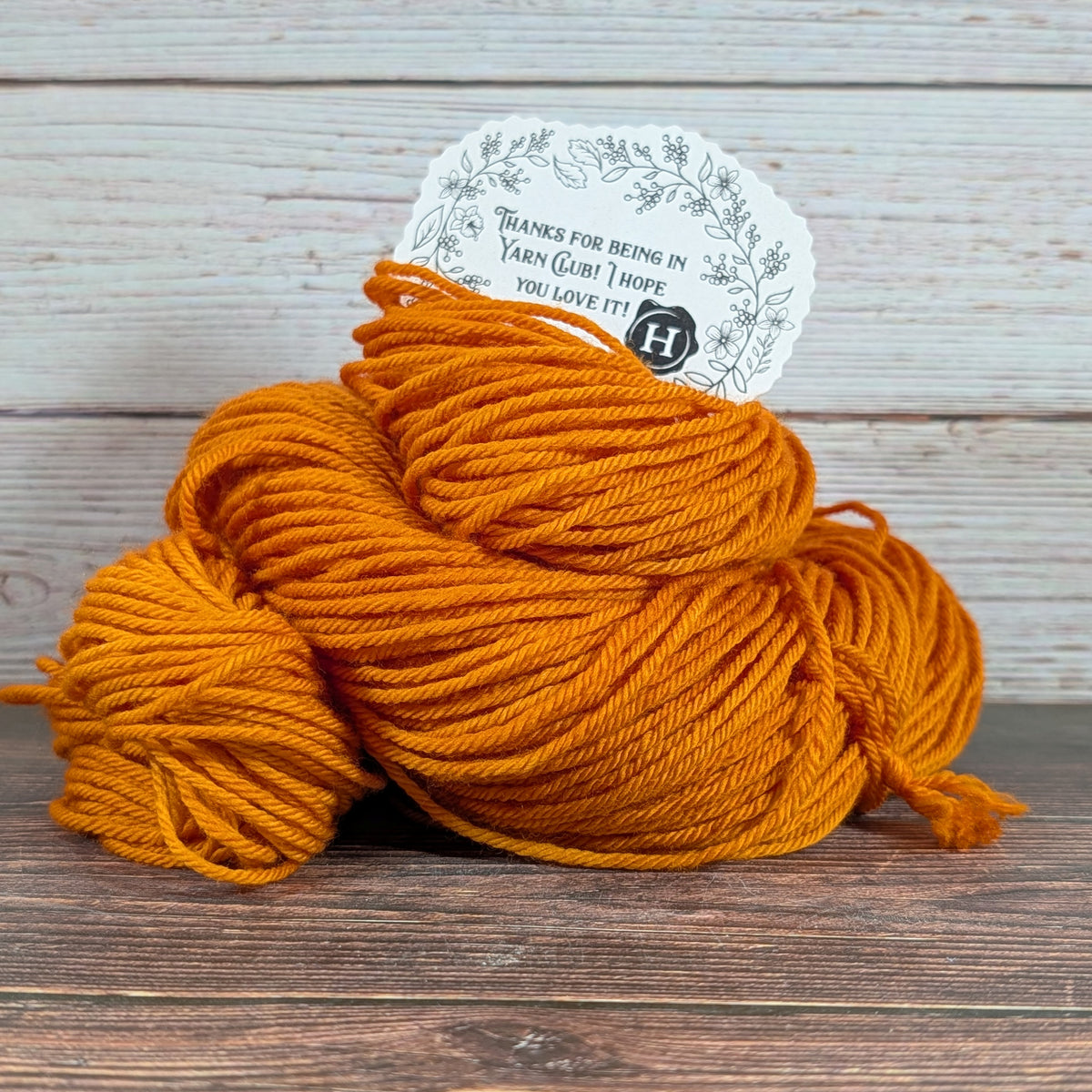 A skein of bright orange yarn with a white paper tag, placed on a dark wooden surface against a light wood plank background.