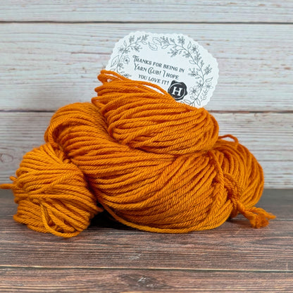 A skein of bright orange yarn with a white paper tag, placed on a dark wooden surface against a light wood plank background.