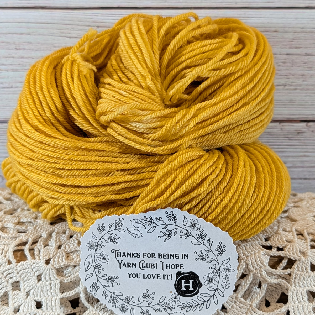 A skein of mustard yellow yarn resting on a cream lace doily against a whitewashed wood plank background, with a white decorative thank-you tag placed in front.