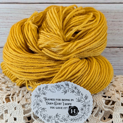 A skein of mustard yellow yarn resting on a cream lace doily against a whitewashed wood plank background, with a white decorative thank-you tag placed in front.