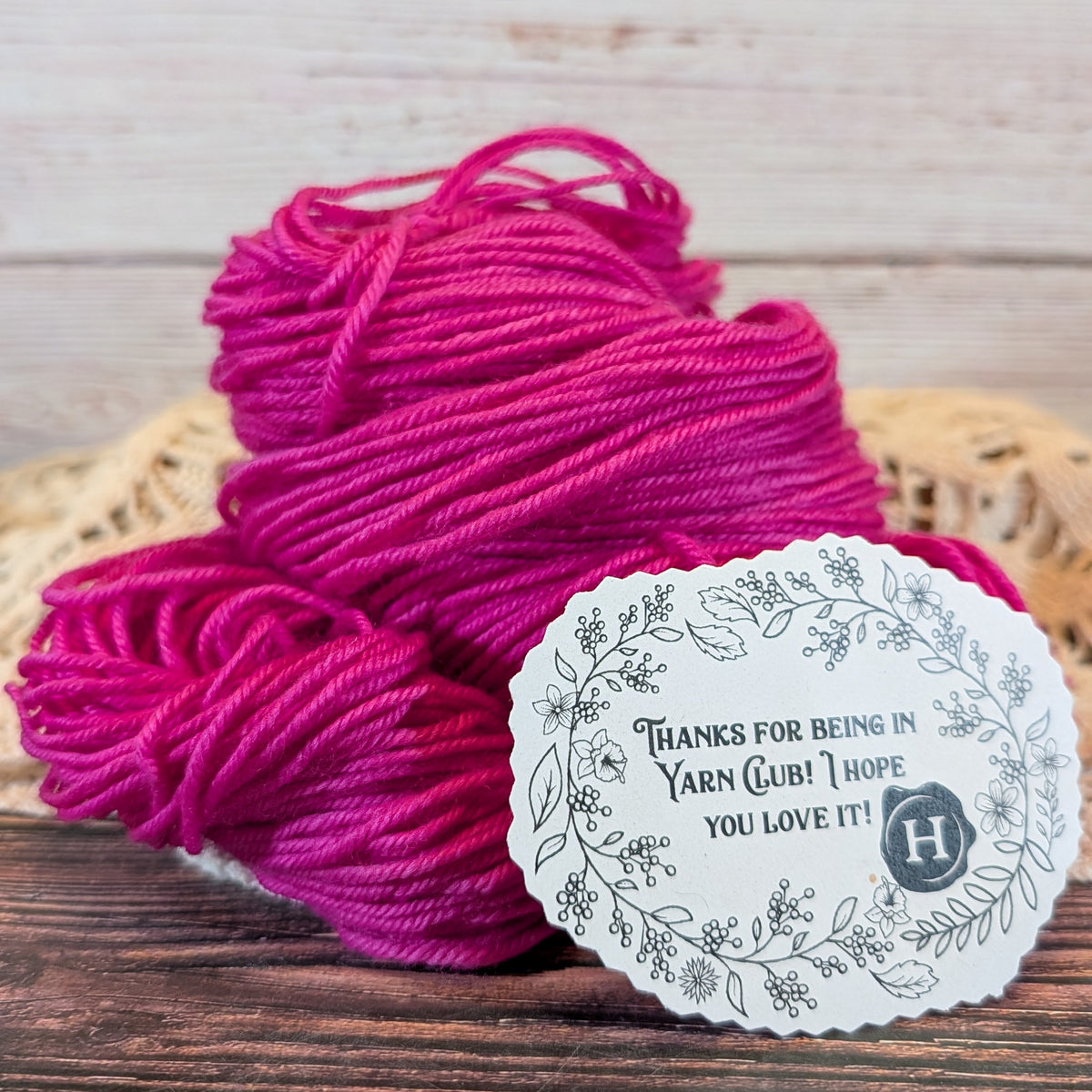 Two skeins of bright magenta pink yarn on a rustic wooden surface with a woven mat underneath and a round white paper tag that reads 'THANKS FOR BEING IN YARN CLUB! I HOPE YOU LOVE IT!!' with a floral border and monogram.