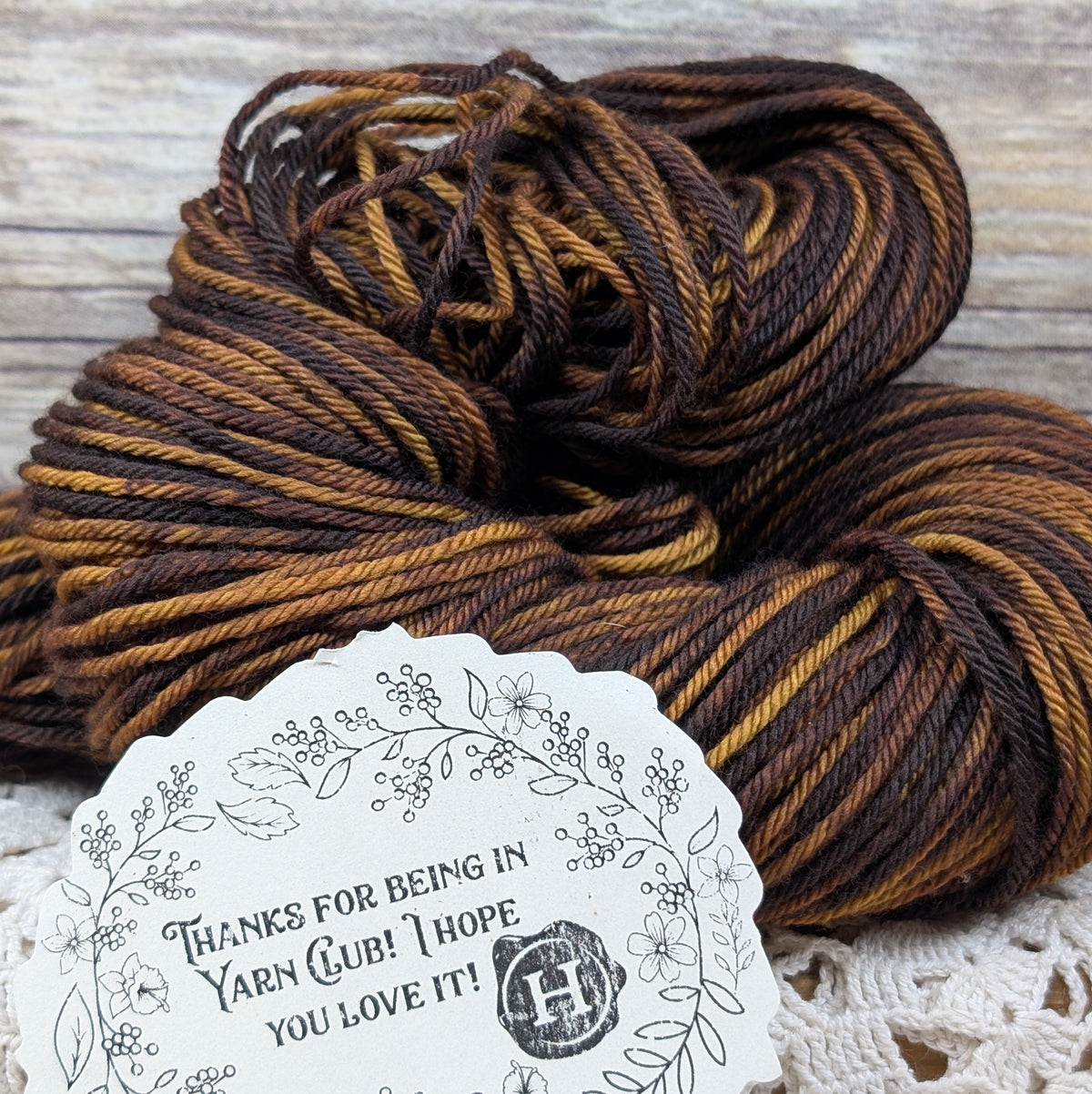 A close-up of a skein of variegated yarn in dark brown and golden tan stripes, with a white scalloped thank-you card placed in front on a rustic wood surface.