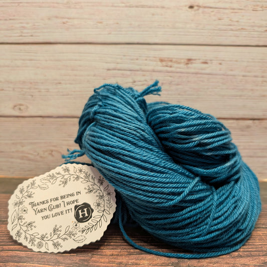 A skein of teal blue yarn resting on a wooden surface next to a round paper tag that reads 'THANKS FOR BEING IN YARN CLUB! I HOPE YOU LOVE IT!'. The background consists of light-colored wooden planks.