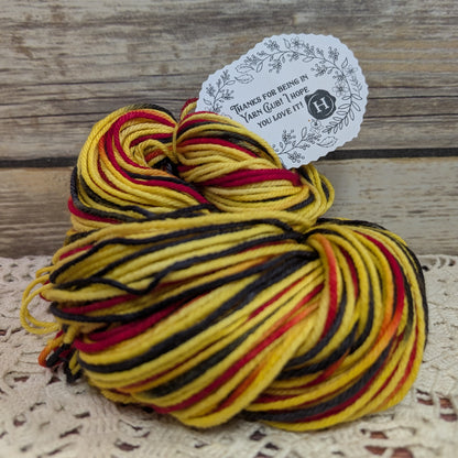 A skein of variegated yarn with yellow, black, and red stripes, featuring a white paper tag with a floral design and text, set on a rustic wooden surface with a lace cloth underneath.