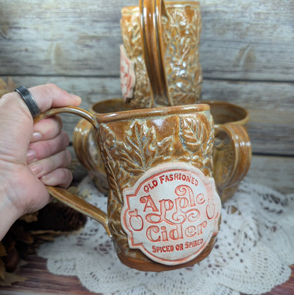 Ceramic mug with 'Old Fashioned Apple Cider' label held by a hand on a wooden surface.