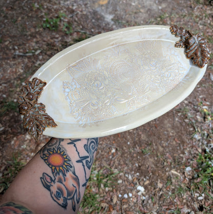 Decorative white oval tray with animal and food patterns held by a person with tattoos.