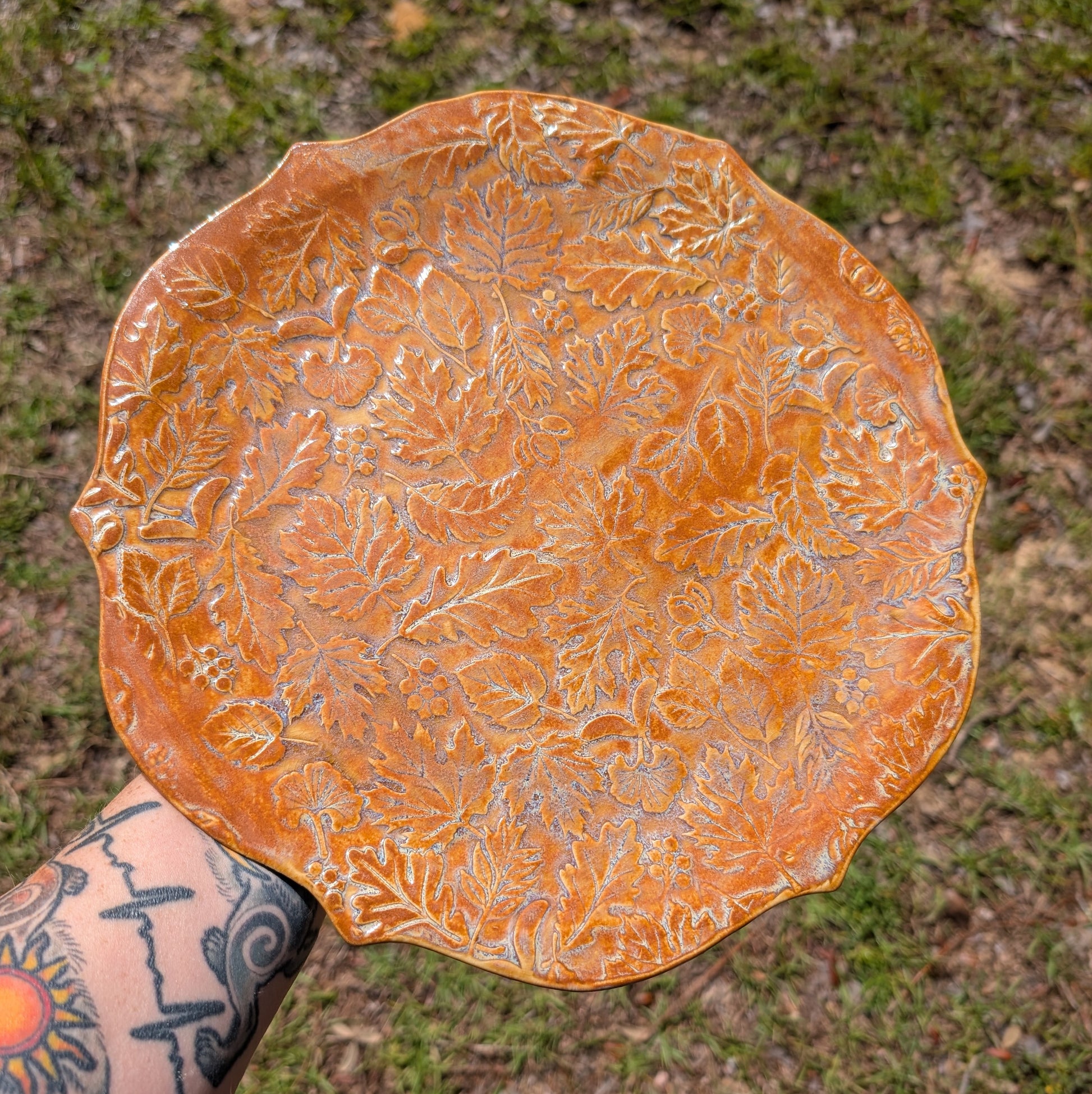 Ceramic plate with leaf patterns held by a person with tattoos, against a natural background.