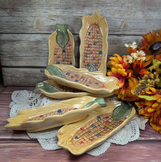 Long thin dishes designed to hold corn on the cob, painted to look like Indian corn on a wooden surface with flowers off to the side. 