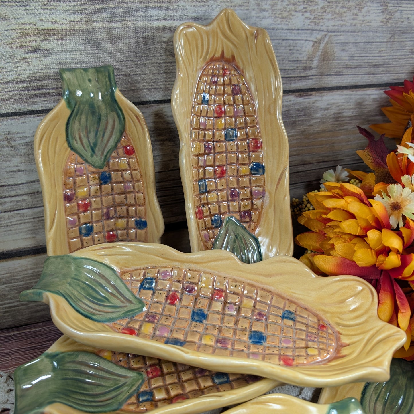 Long thin dishes designed to hold corn on the cob, painted to look like Indian corn on a wooden surface with flowers off to the side. 