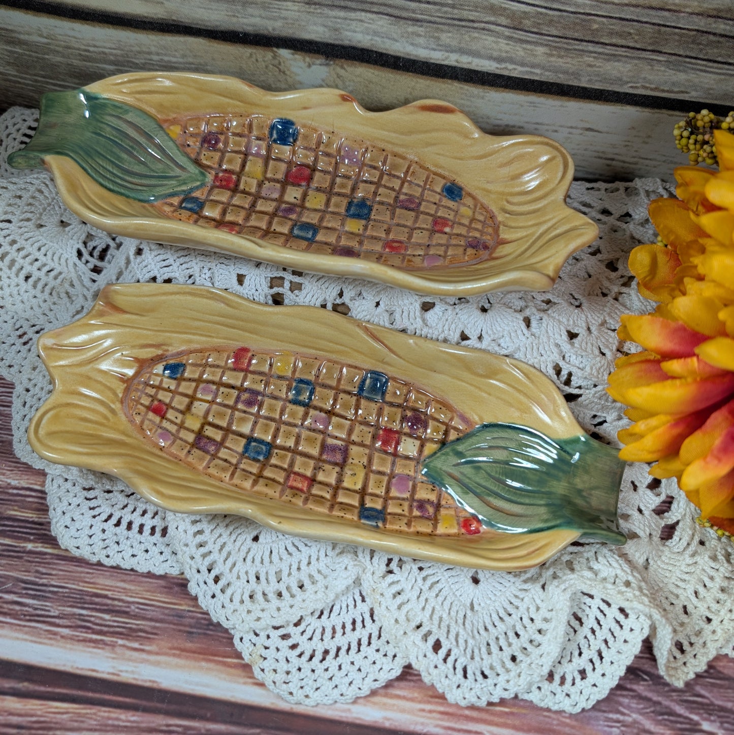 Two dishes the size, shape, and designed like an ear of corn on a lace doily on a wooden background. 