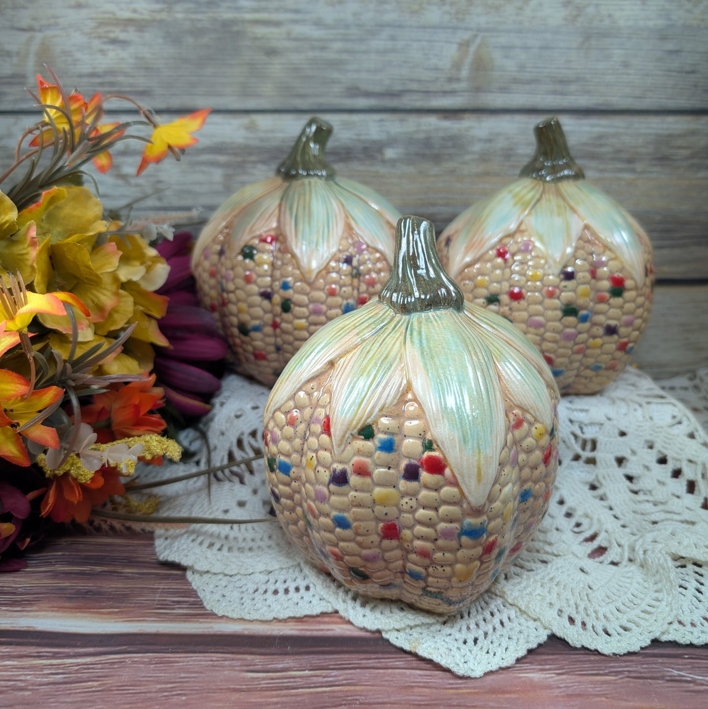 Decorative corn and pumpkin figurines on a lace doily with flowers and wooden background