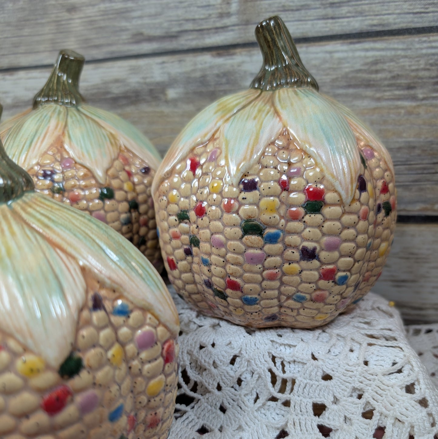 Decorative corn and pumpkin-shaped ceramic pieces on a lace tablecloth with a wooden background.