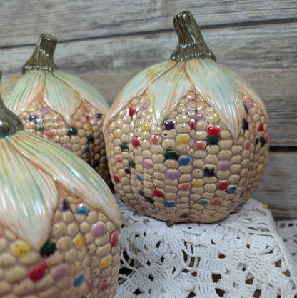 Decorative corn and pumpkin-shaped ceramic pieces on a lace tablecloth with a wooden background.