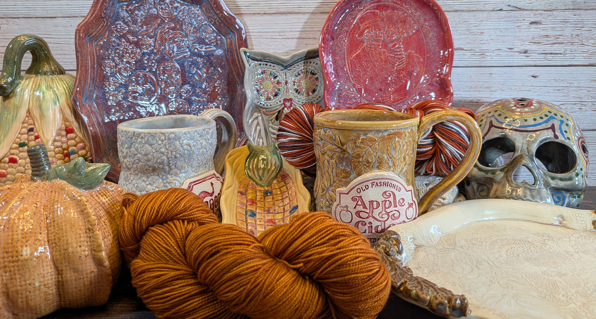 A collection of autumn-themed ceramic tableware and decor, including mugs, plates, a pumpkin-shaped vase, a skull, an owl figurine, and skeins of orange yarn, arranged against a rustic white wood plank background.