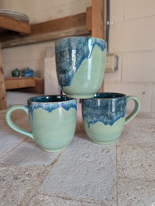Three ceramic mugs with blue and green glaze stacked on a stone surface.