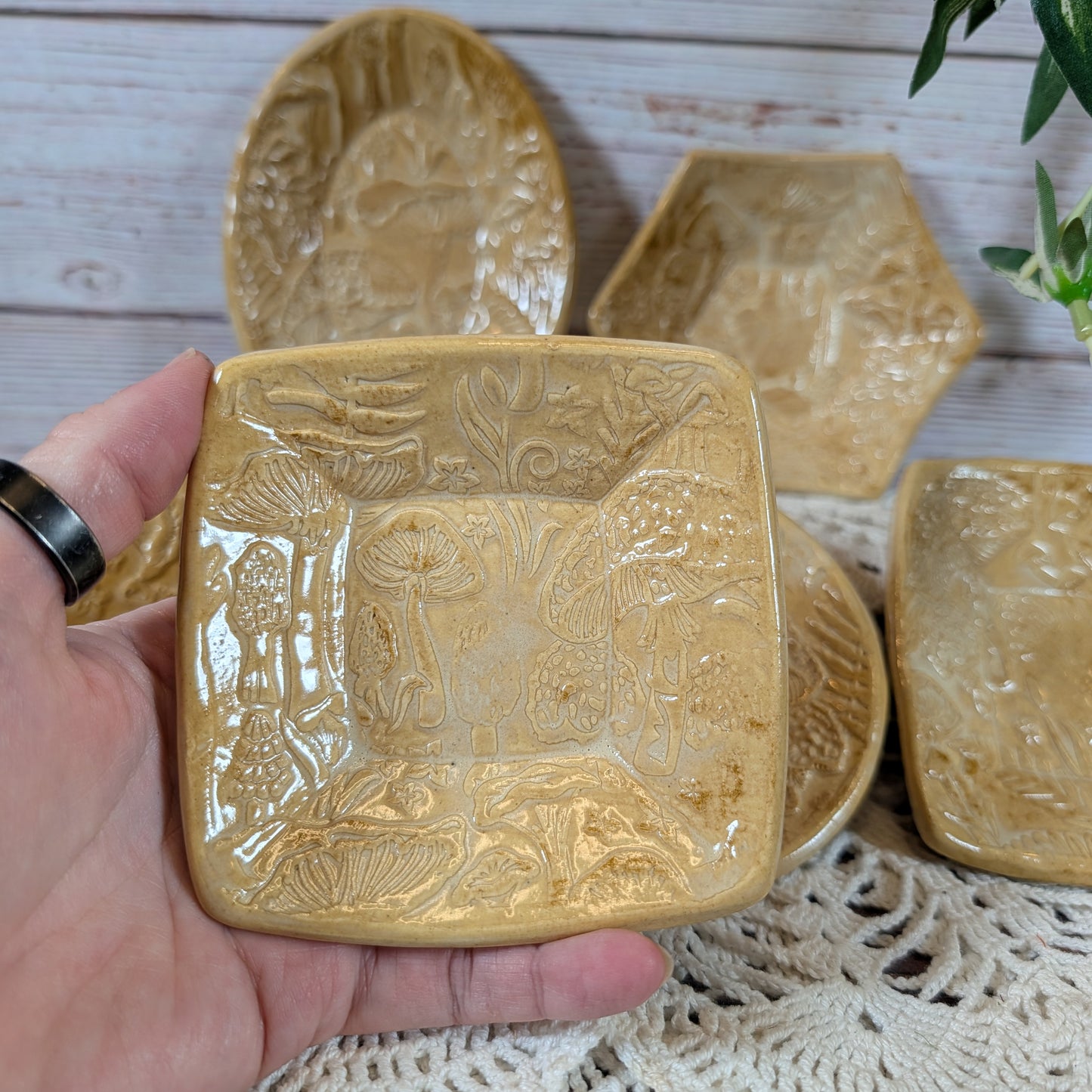 A hand holding a small square dish. In back is the rest of the set of small ceramic trinket dishes, decorated with a mushroom motif, on a lace doily with a wooden background