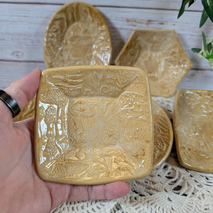 A hand holding a small square dish. In back is the rest of the set of small ceramic trinket dishes, decorated with a mushroom motif, on a lace doily with a wooden background