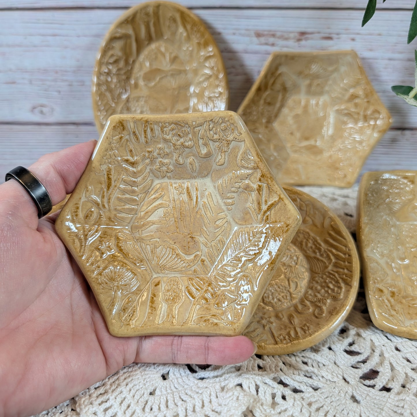 A hand holding a small hexagonal dish. In back is the rest of the set of small ceramic trinket dishes, decorated with a mushroom motif, on a lace doily with a wooden background