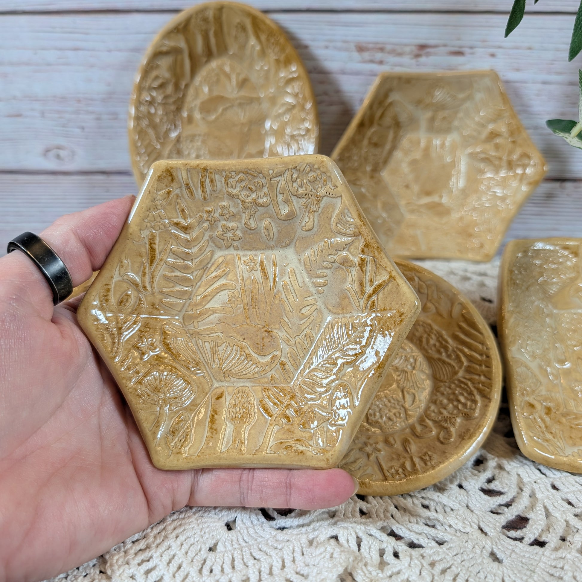 A hand holding a small hexagonal dish. In back is the rest of the set of small ceramic trinket dishes, decorated with a mushroom motif, on a lace doily with a wooden background