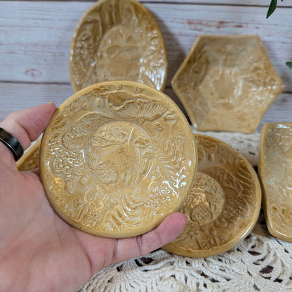 A hand holding a small round dish. In back is the rest of the set of small ceramic trinket dishes, decorated with a mushroom motif, on a lace doily with a wooden background