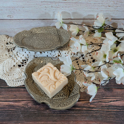 A rectangular cream-colored soap bar with an embossed floral pattern sits on an ornate oval ceramic soap dish. Another matching dish is behind it, and white blossoms are arranged on a lace doily over a rustic wooden surface.