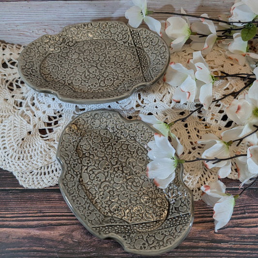 Two scalloped ceramic soap dishes with embossed patterns in a muted gray glaze, styled on a white lace doily over a dark wood surface with white blossoms.
