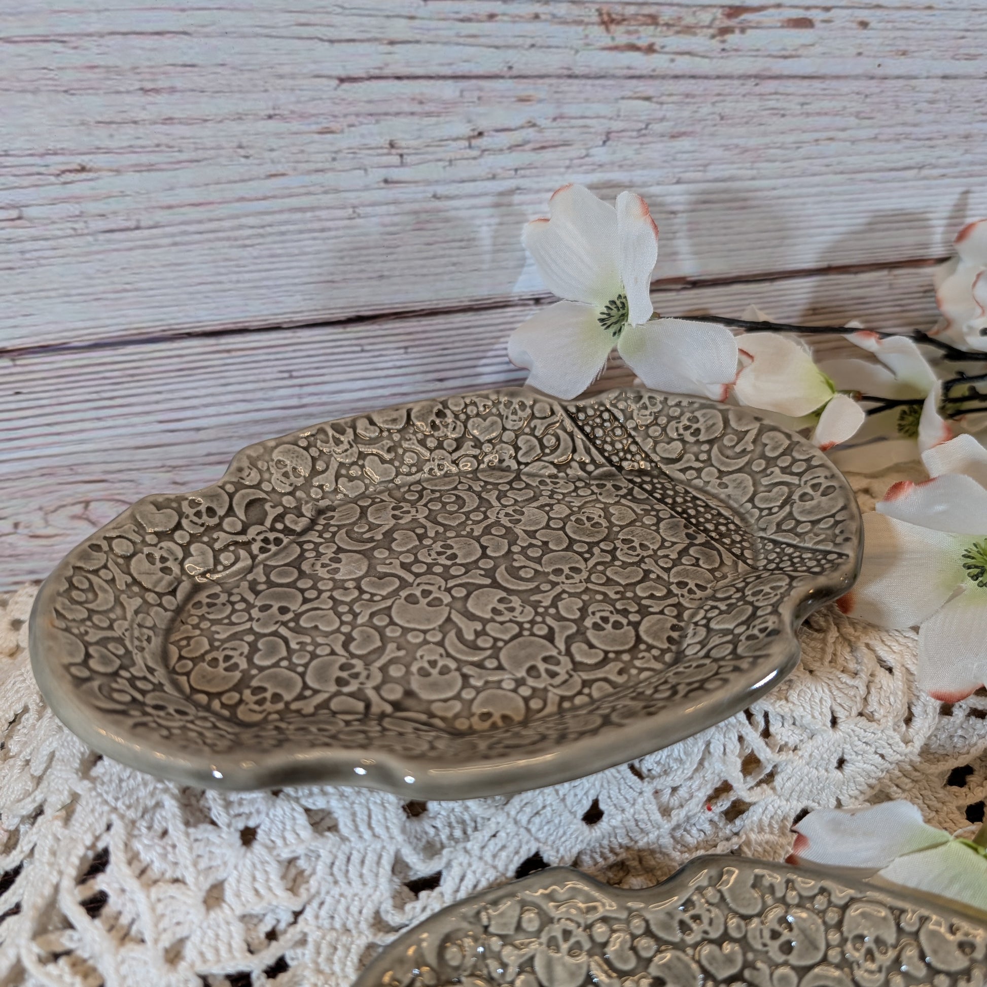 An oval soap dish with an embossed pattern in a muted gray glaze, styled on a white crocheted doily with white flowers against a rustic whitewashed wood background.