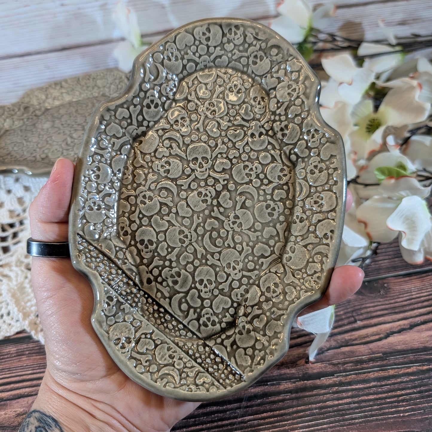 A hand holds a decorative ceramic soap dish with an embossed skull pattern, set against a rustic wooden background with white flowers.