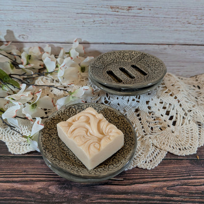 A rectangular bar of cream-colored soap with a carved swirl pattern sits on a round textured dish, with a matching soap dish lid behind it on a lace doily over a wooden surface.