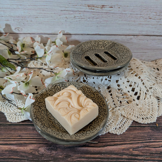 A rectangular bar of cream-colored soap with a carved swirl pattern sits on a round textured dish, with a matching soap dish lid behind it on a lace doily over a wooden surface.
