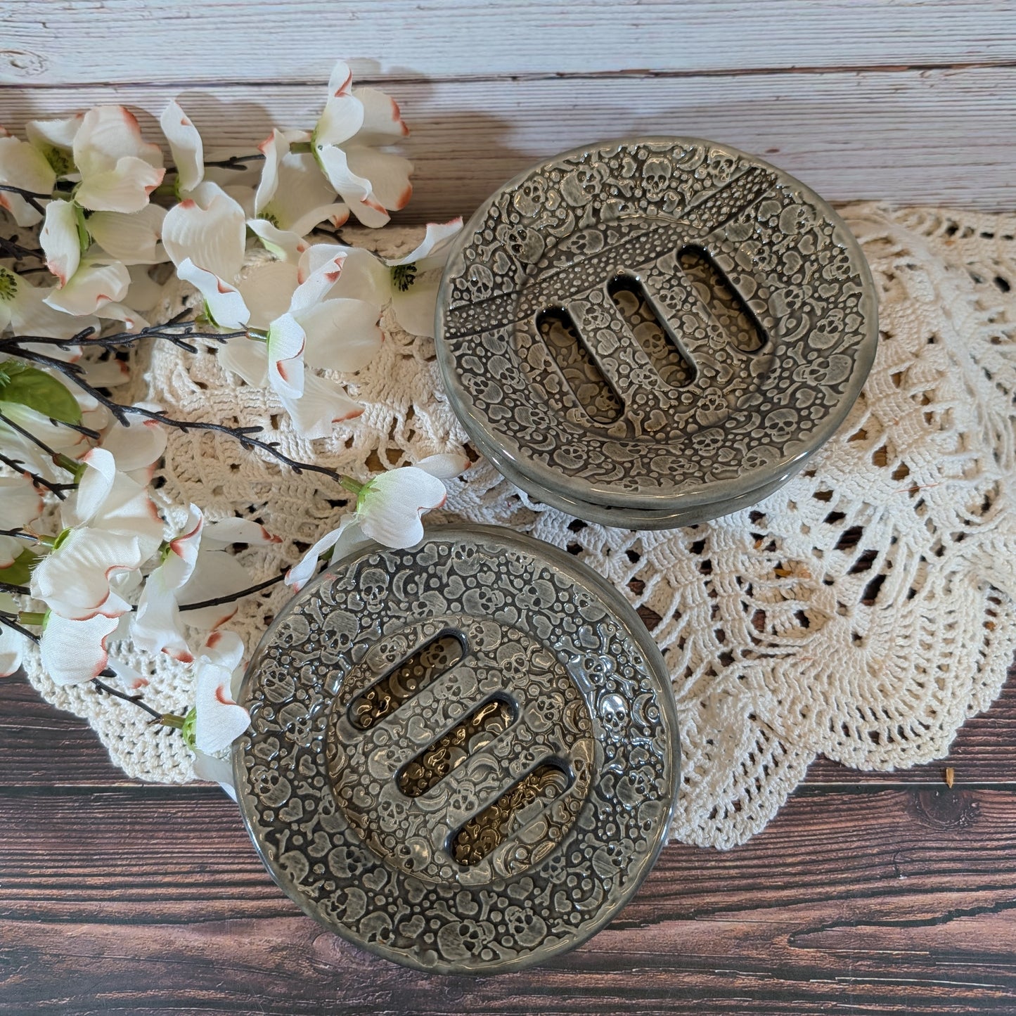 Two round ceramic soap dishes with a carved floral pattern and three slotted centers, displayed on a lace doily over a wooden surface with white flowers.