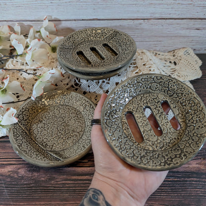 Three round ceramic soap dish soakers with intricate floral embossing and drainage slots, shown on a rustic wooden table with a hand holding one in the foreground.