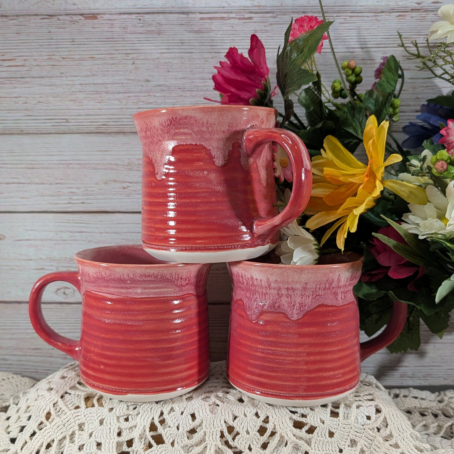 Three handmade ceramic mugs in a coral-pink and red glaze, stacked in a pyramid on a white lace doily against a rustic white wood plank background with a flower arrangement.