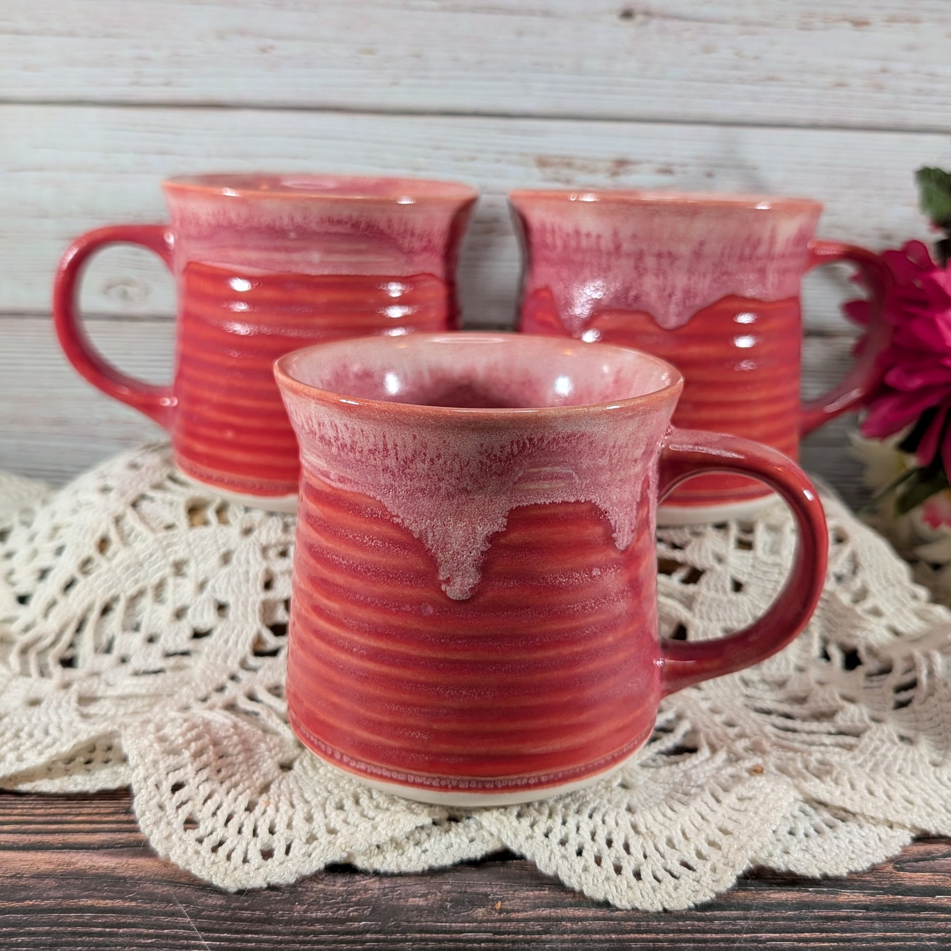Three handmade ceramic mugs with a glossy red and pink dripping glaze, arranged on a white crocheted doily over a rustic wooden table.