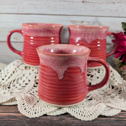 Three handmade ceramic mugs with a glossy red and pink dripping glaze, arranged on a white crocheted doily over a rustic wooden table.