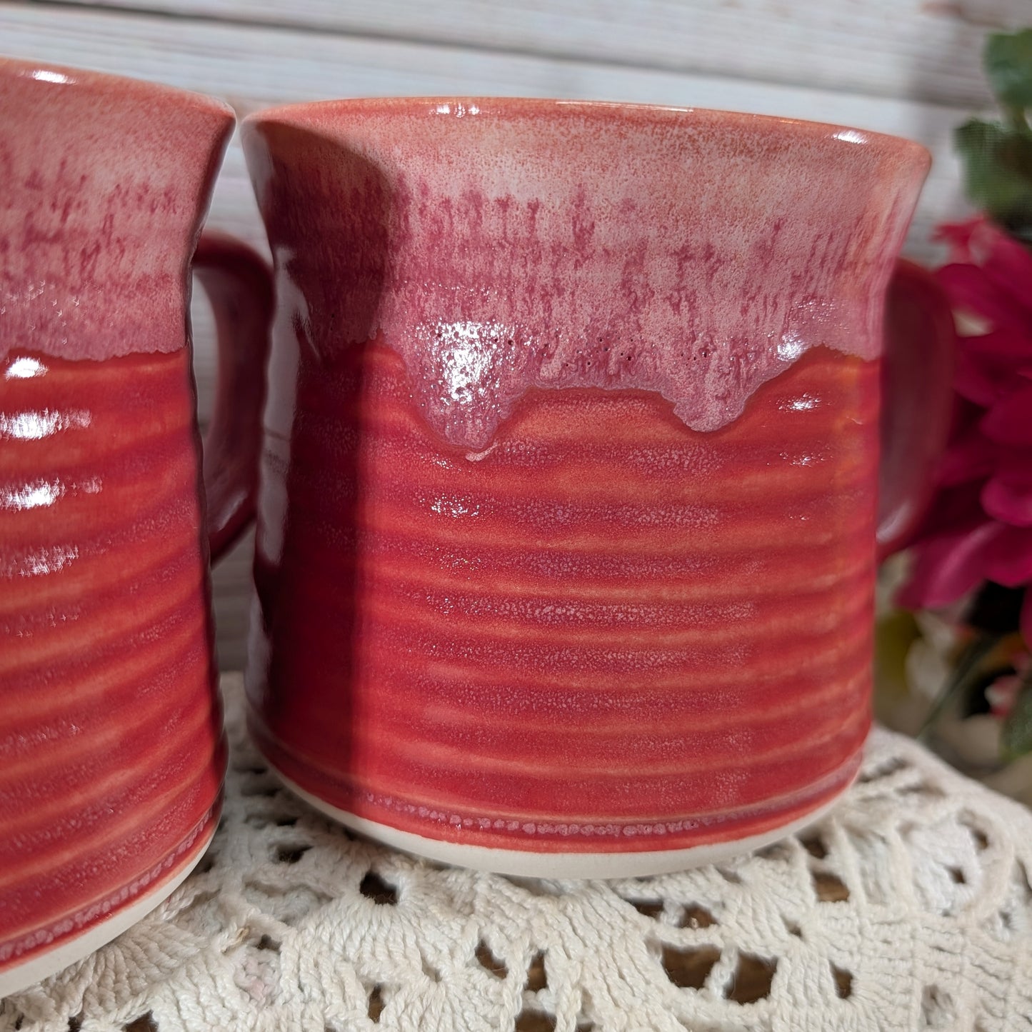 A close-up of a handmade ceramic mug with a glossy dripping pink and red glaze, resting on a white lace doily with another mug partially visible on the left.