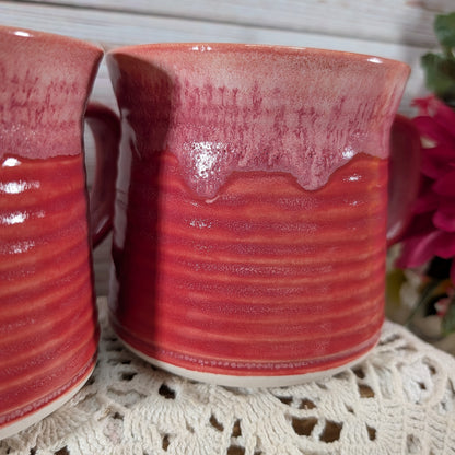 A close-up of a handmade ceramic mug with a glossy dripping pink and red glaze, resting on a white lace doily with another mug partially visible on the left.